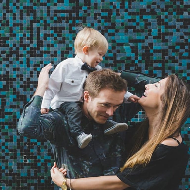 Family playing in front of a blue wall. The little boy is sitting on the dads shoulders