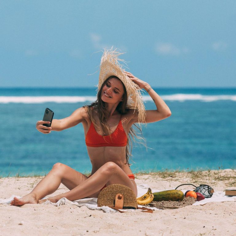Woman on the beach wearing a red bathing suit and straw hat taking a selfie