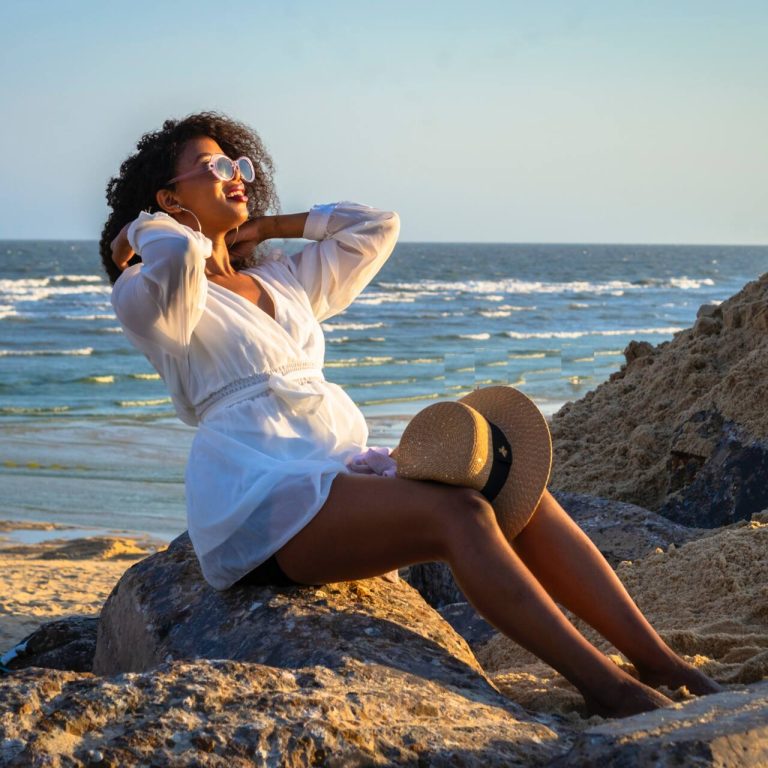 Woman in a white cover up on the beach sitting on a rock